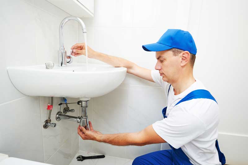 A man in blue overalls repairing a sink in a kitchen.