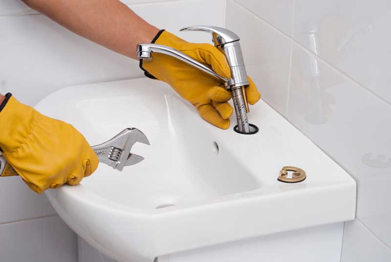 A plumber with yellow gloves installing a sink faucet using a wrench.