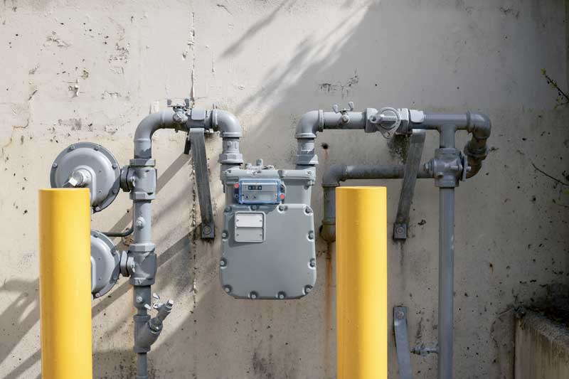 A gas meter mounted on a brick wall, with a yellow gas line entering the meter on the left and a yellow gas line exiting the meter on the right.